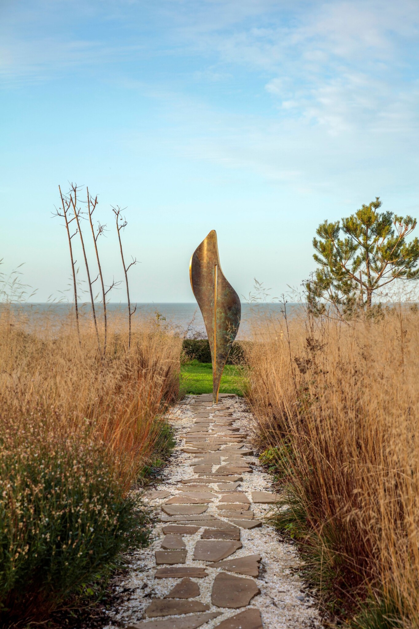 A stone path winds through tall golden grass to a contemporary abstract sculpture made from bronze finish and gold leaf, shaped like a vertical leaf. Green grass, a tree, and the calm blue sea sit beneath a partly cloudy sky.