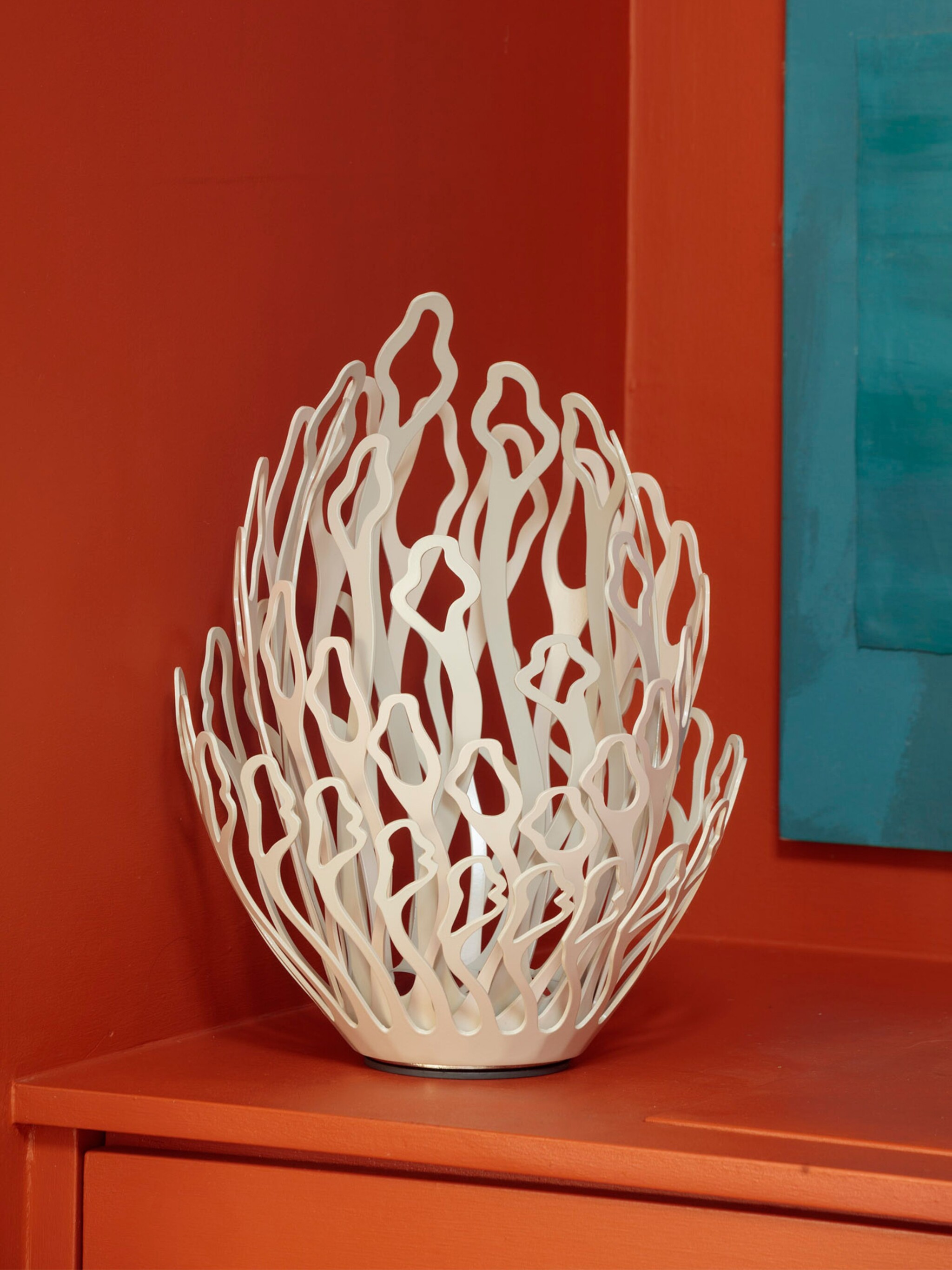 A decorative white bowl with an open, abstract, coral-like design sits on an orange surface against an orange wall. Part of a teal artwork is visible on the wall to the right, adding contrast to the composition.