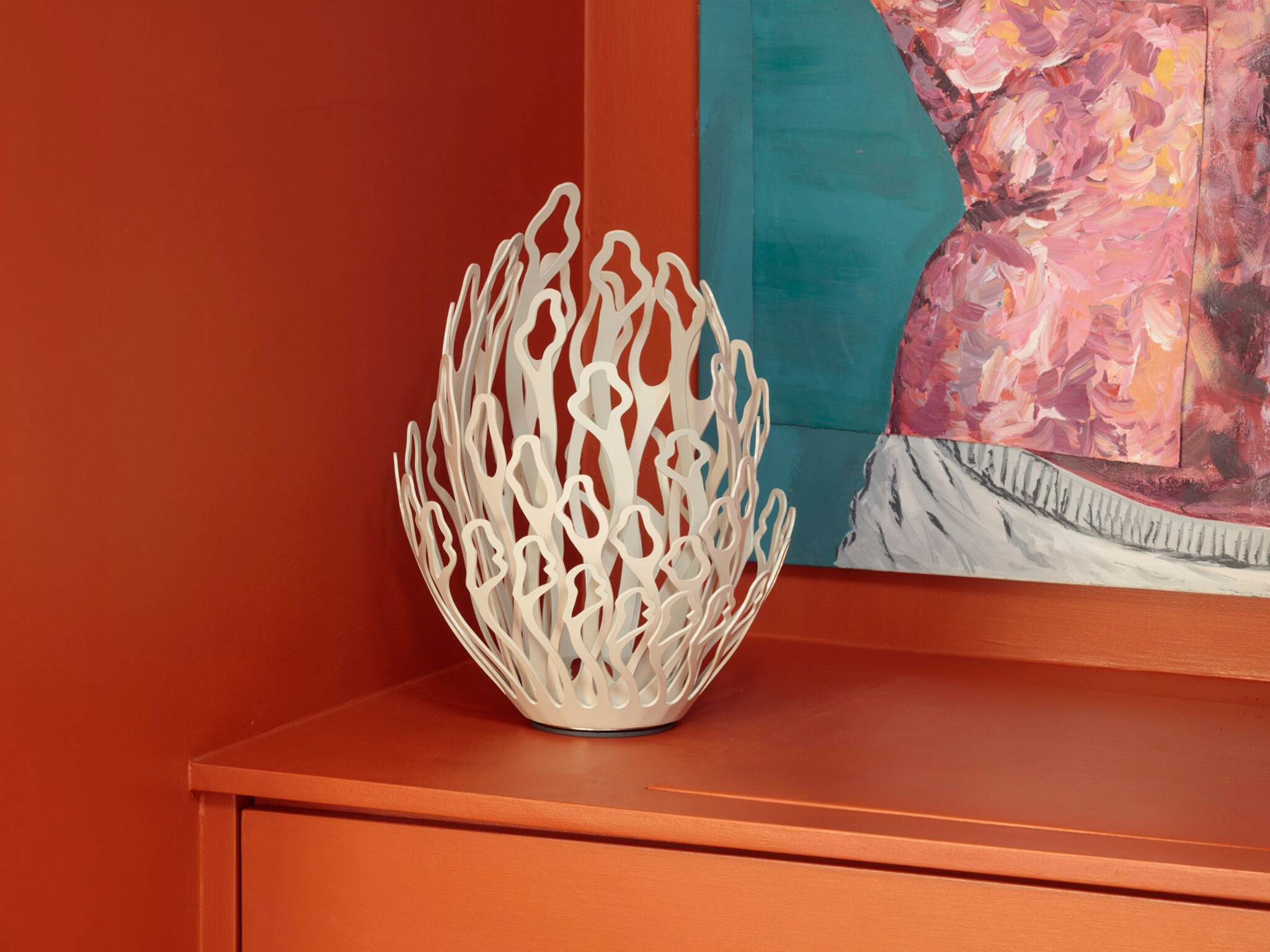 A white metal sculpture with an open, geometric design sits on an orange surface against an orange wall. Part of an abstract painting with pink, blue, and grey brushstrokes is visible in the background.
