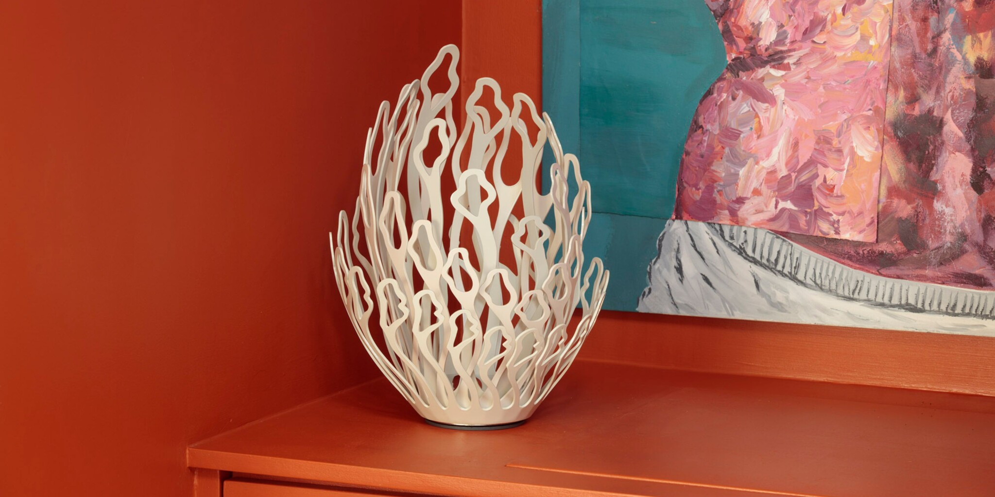 A white, abstract, coral-like decorative sculpture sits on an orange surface in the corner of a room with orange walls. Part of a colourful painting with pink, teal, and white tones is visible on the wall behind it.