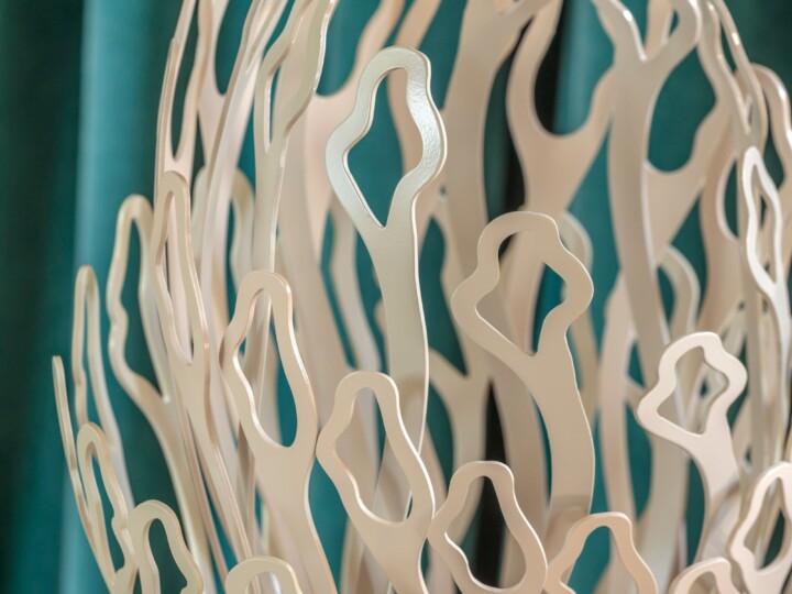 Close-up of an abstract metal sculpture featuring interwoven, elongated shapes with open, rounded cut-outs. The sculpture is cream-coloured and set against a soft-focus teal background, giving a sense of flow and organic movement.