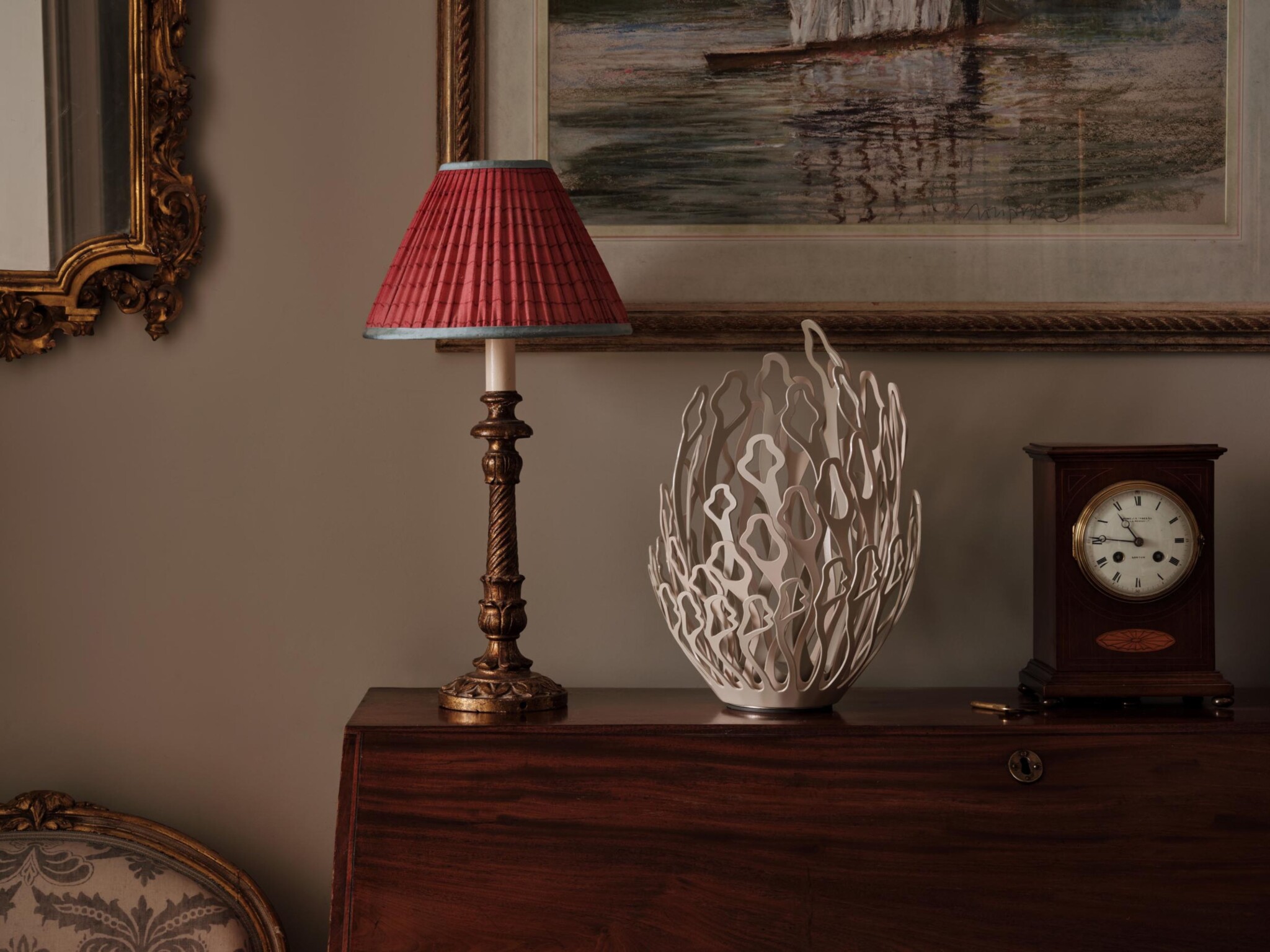A classic room scene with a gilt-framed mirror, a red pleated lampshade on a bronze lamp, a modern white ceramic sculpture, a dark wood mantel clock with a white face, and a framed painting above a polished wooden surface.
