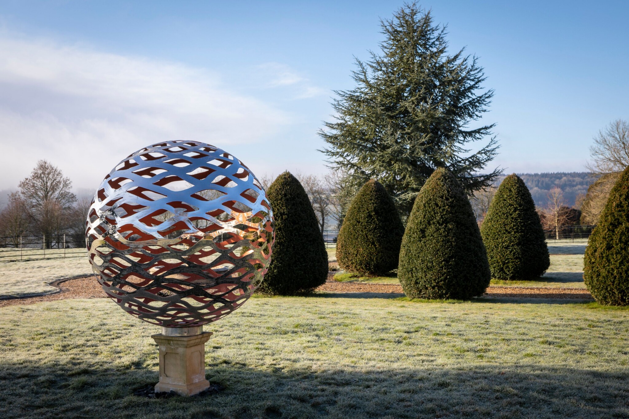 A large metallic sphere sculpture stands on a plinth in a landscaped garden with trimmed shrubs.