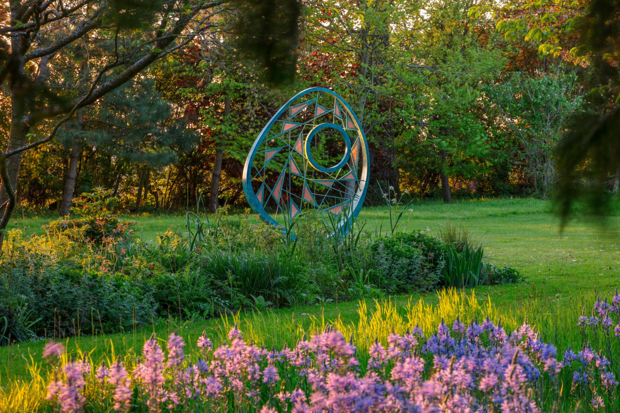 David Harber woven bronze sculpture, Nexa, is placed on a lush green lawn surrounded by leafy trees to provide a focal point for bedding that houses late season tulips and cammassia.