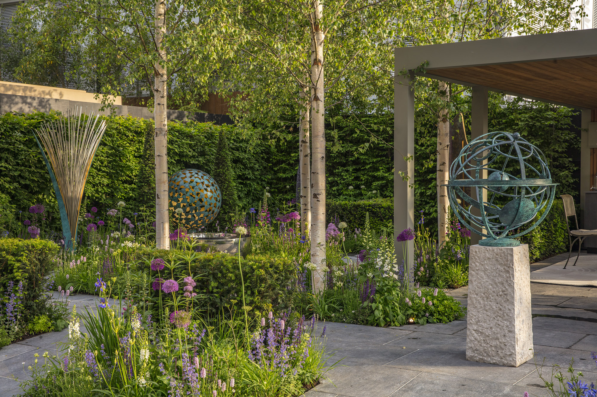 A modern garden with sculptures, trees, flowers, and a shaded seating area under a wooden canopy.