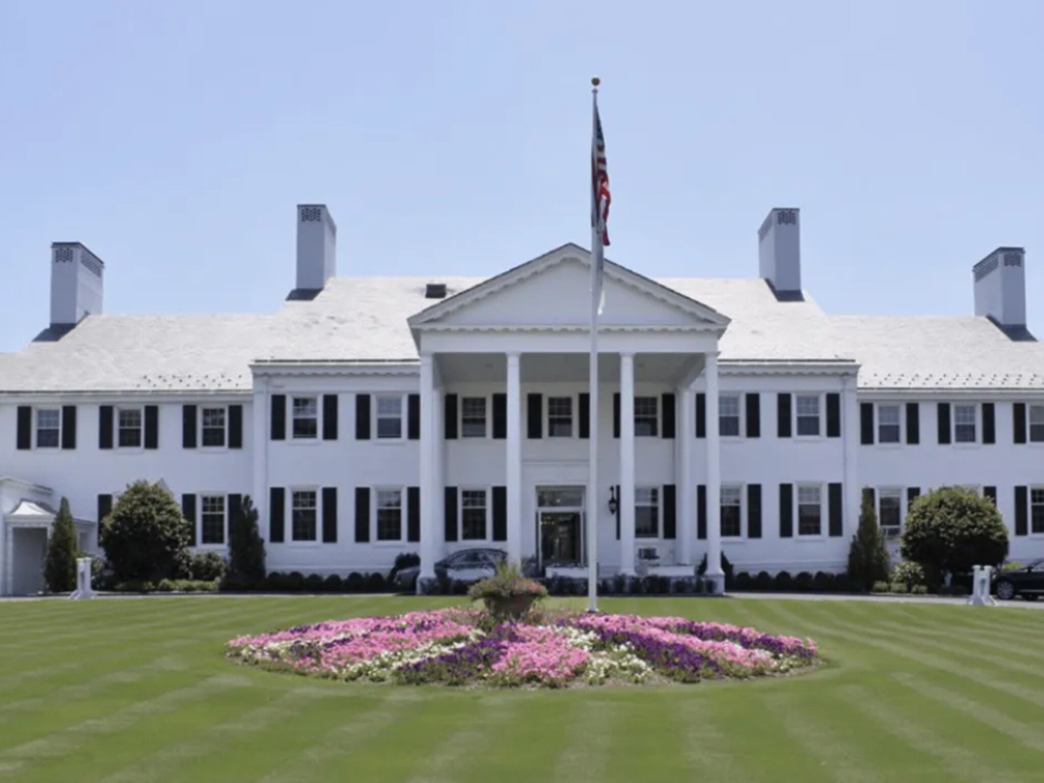 A large, white, two-storey mansion with tall columns and black shutters stands behind a manicured lawn—perfect for hosting inspiring events like Designing Greenwich 2025. A circular flower bed and a flagpole with an American flag enhance the grand setting.
