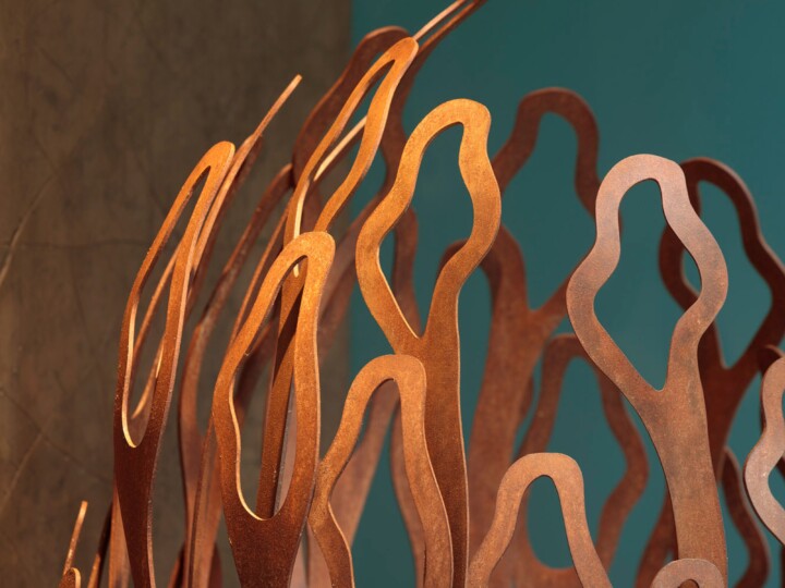 Curved, abstract metal shapes in a warm copper colour rise upward, resembling organic forms or stylised flames. The background transitions from a teal wall on the right to a brownish surface on the left, creating a striking contrast with the sculpture.