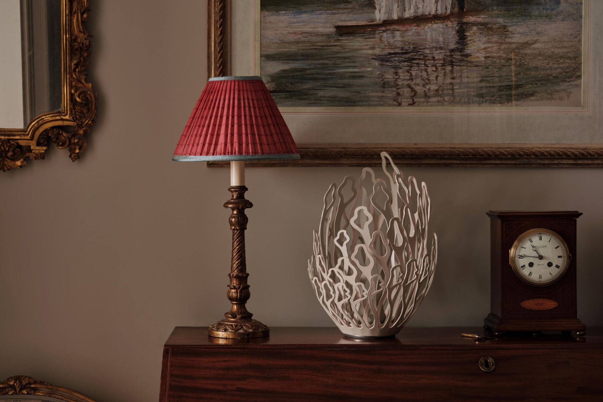 A warmly lit interior scene shows a wooden chest of drawers topped with a brass lamp with a red pleated shade, an ornate white sculptural vase, and a vintage wooden clock. A framed painting with a sailing boat hangs on the beige wall above.