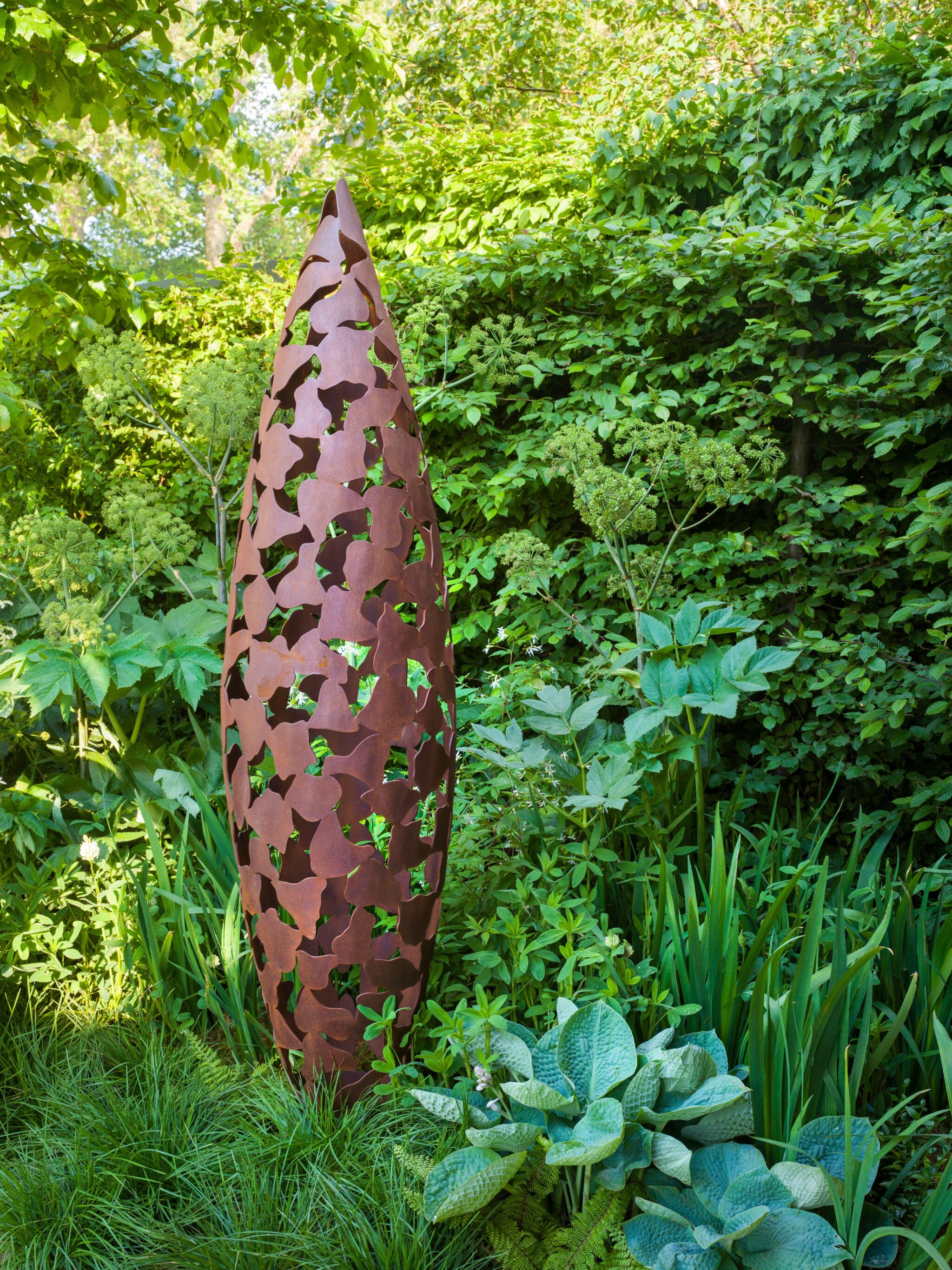 Tall sculpture with a rust-like finish, which contrasts with the textural green planting its positioned in