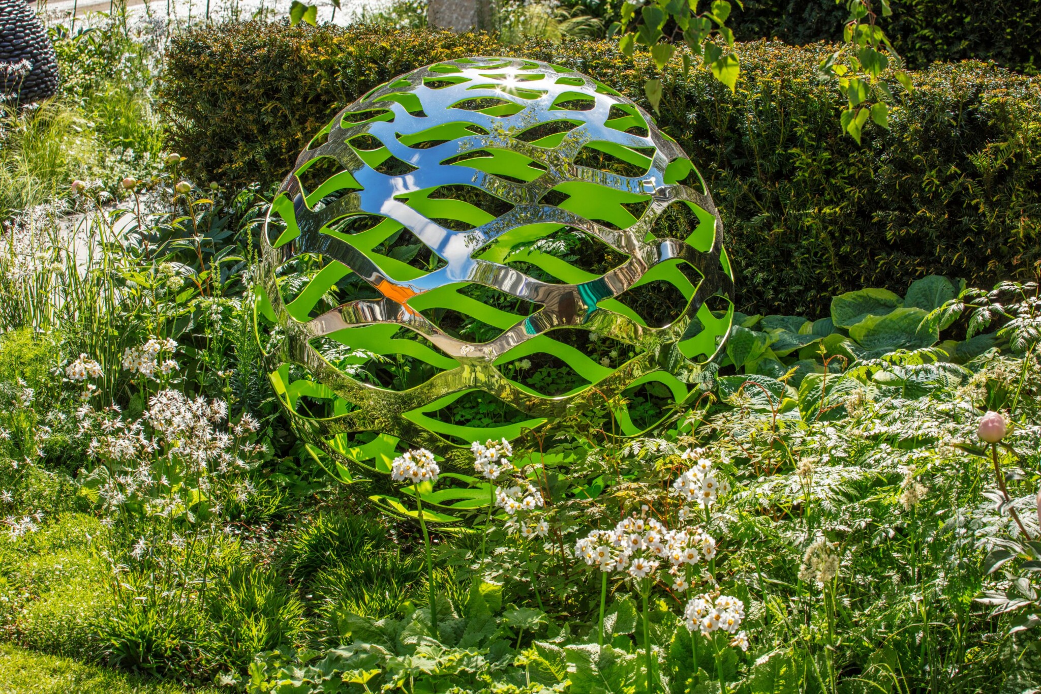 Stainless steel sphere sculpture with green accents sits in green and white spring planting