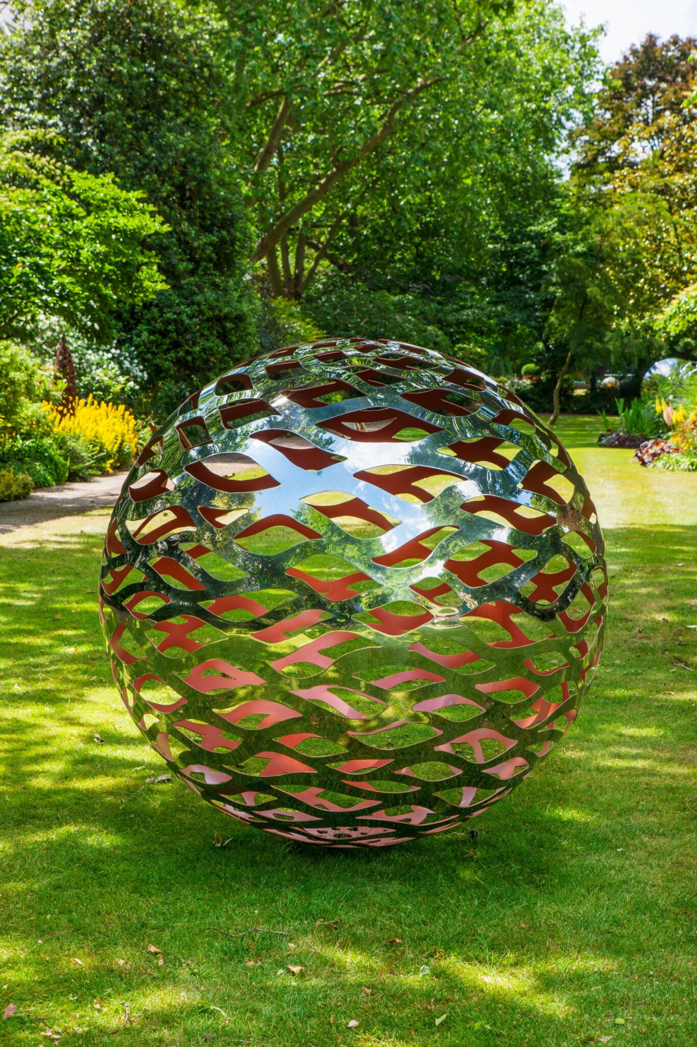 Stainless steel artwork with red hues sits on a manicured lawn with trees in the background.