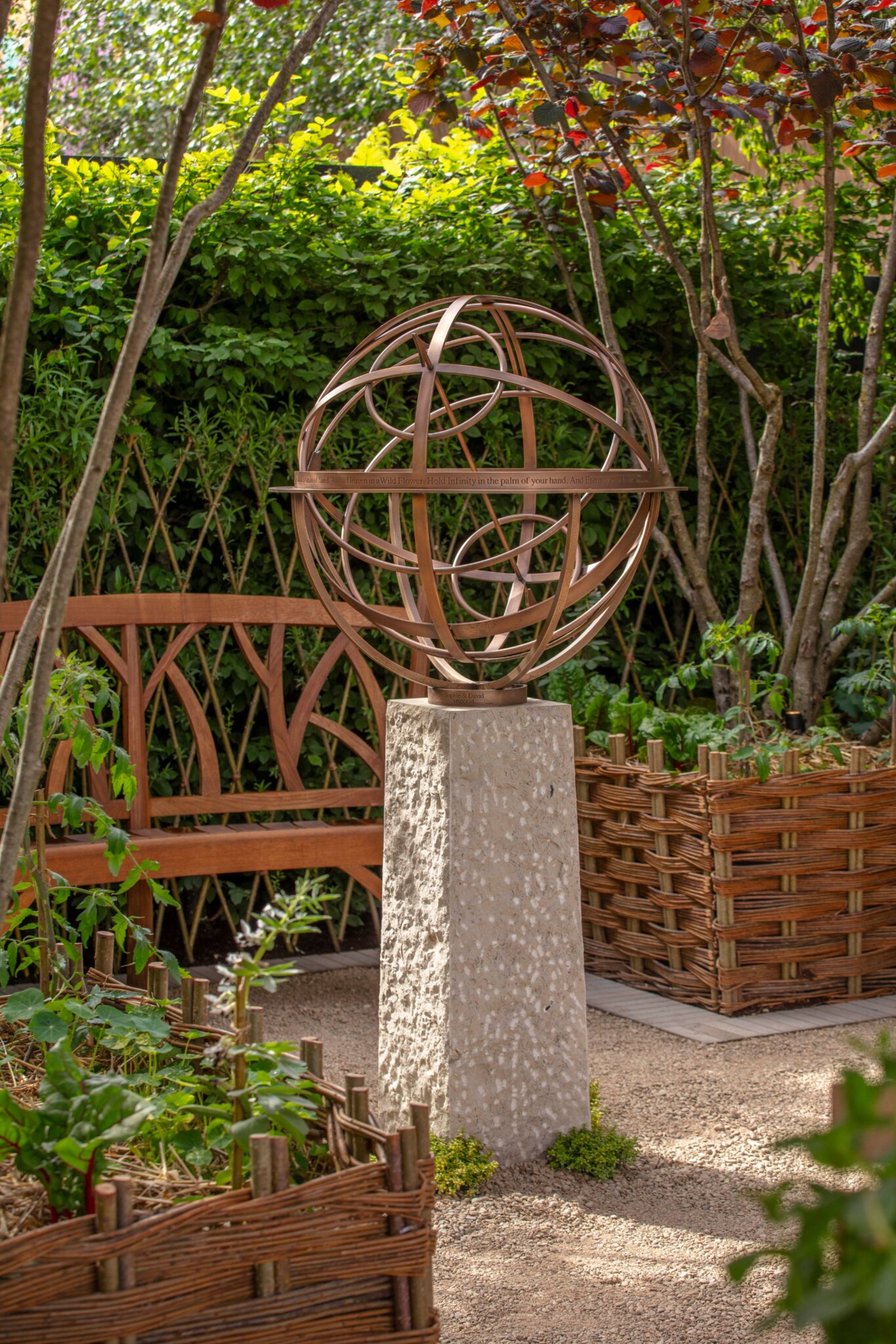 A bronze Armillary Sphere raised onto a plinth sits in a couryard garden space with woven planters and multi-stem trees.