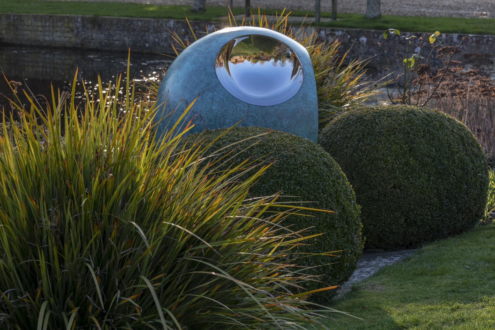 Garden sculpture with a shiny, round reflective surface is an abstract contemporary piece among bushes by the pond.