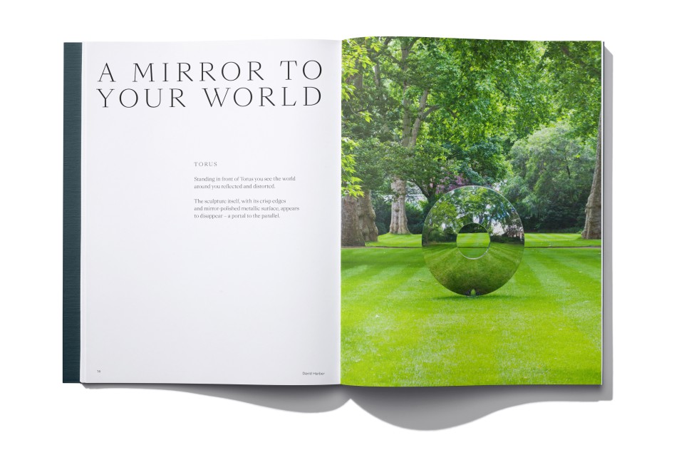 A book spread shows the left page with the title A MIRROR TO YOUR WORLD and text, while the right page features a photo of a shiny stainless steel torus contemporary sculpture on a manicured lawn, reflecting trees and greenery in a garden setting.