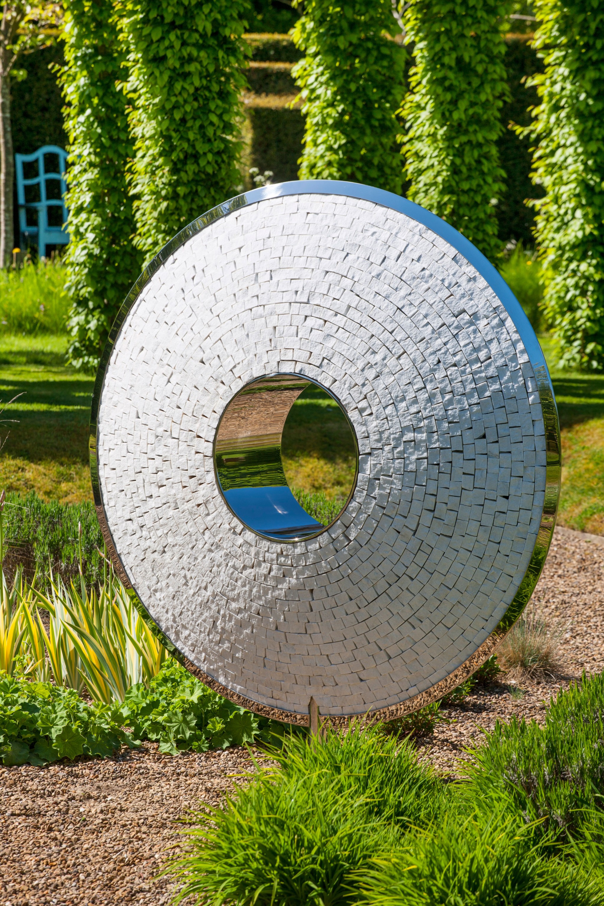 A shiny torus-shaped stainless steel sculpture with a hole stands in a lush green garden.
