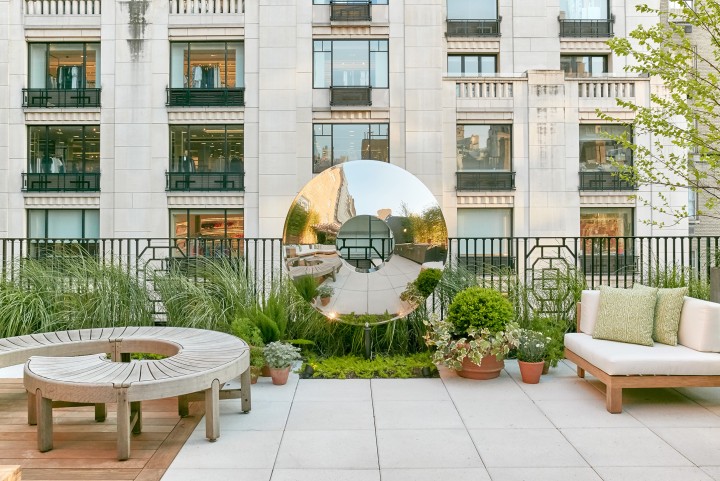A rooftop patio with modern outdoor seating, potted plants, and a large torus stainless steel contemporary abstract sculpture reflecting the surroundings. In the background, a tall stone building with many windows and balconies is visible. Lush greenery borders parts of the patio.