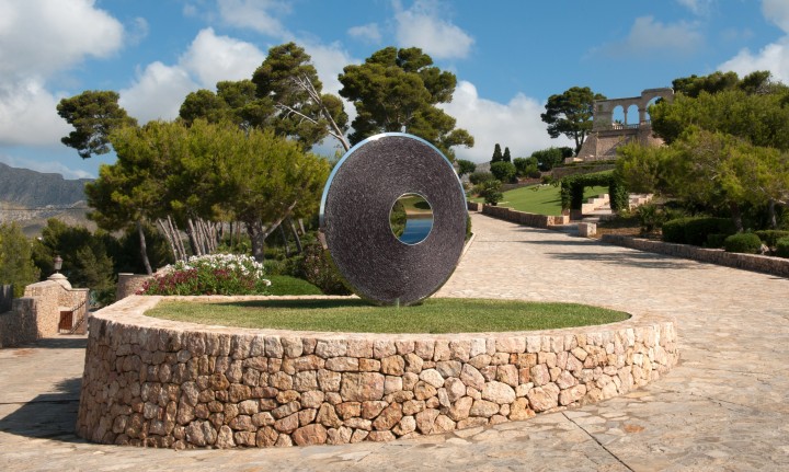 A large, round torus sculpture with a hole in the centre sits on a small grassy area, surrounded by a low stone wall. This contemporary abstract sculpture, made of stainless steel and slate, stands along a wide stone pathway lined with trees.