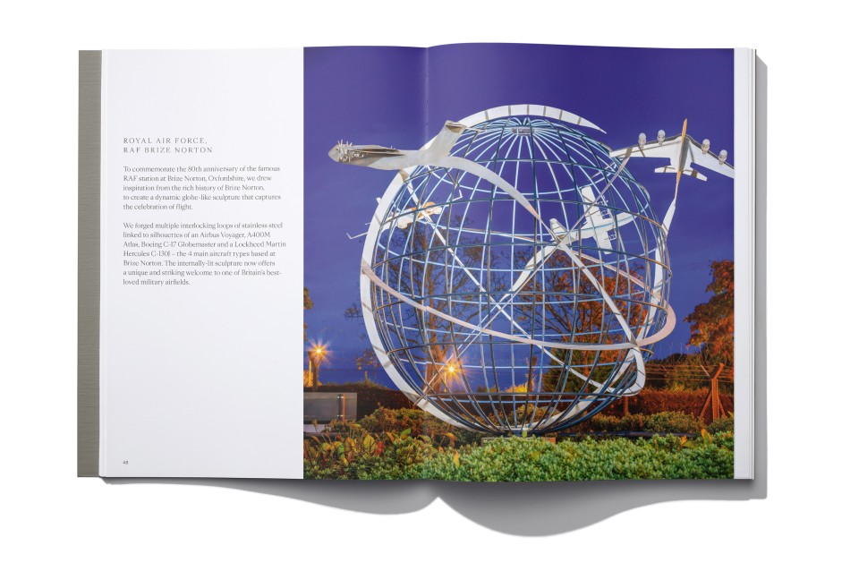 An open book displays a large RAF Brize Norton bespoke globe sculpture with three model aeroplanes mounted on it, set outdoors against a twilight sky. The left page has text about the Royal Air Force; the right shows the sculpture amid greenery and lights.