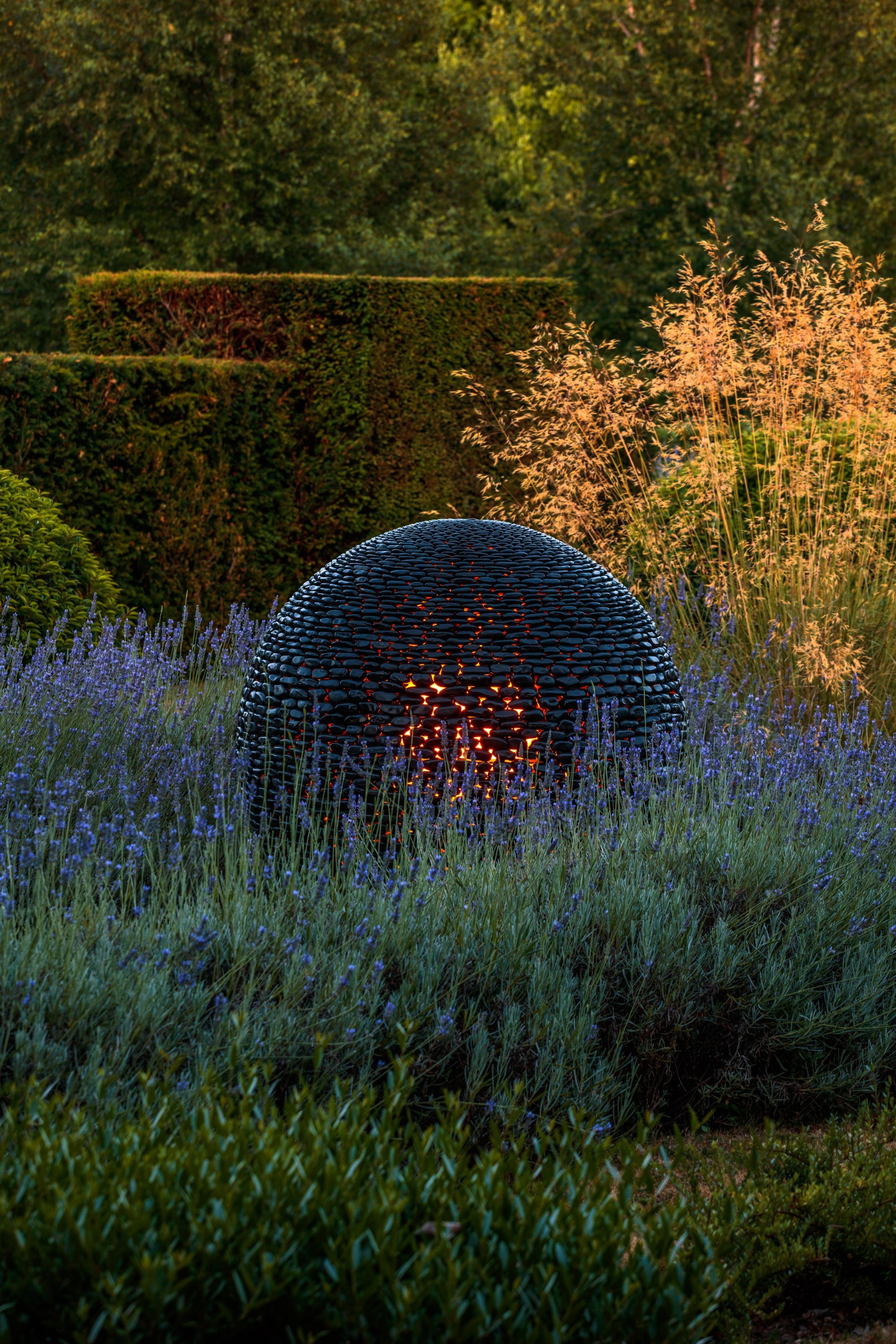 A dark planet stone contemporary sculpture with a honeycomb texture sits among tall grasses and purple flowers in a garden. Warm orange light glows from its hollow centre, framed by lush green hedges and sunlit plants in the background.