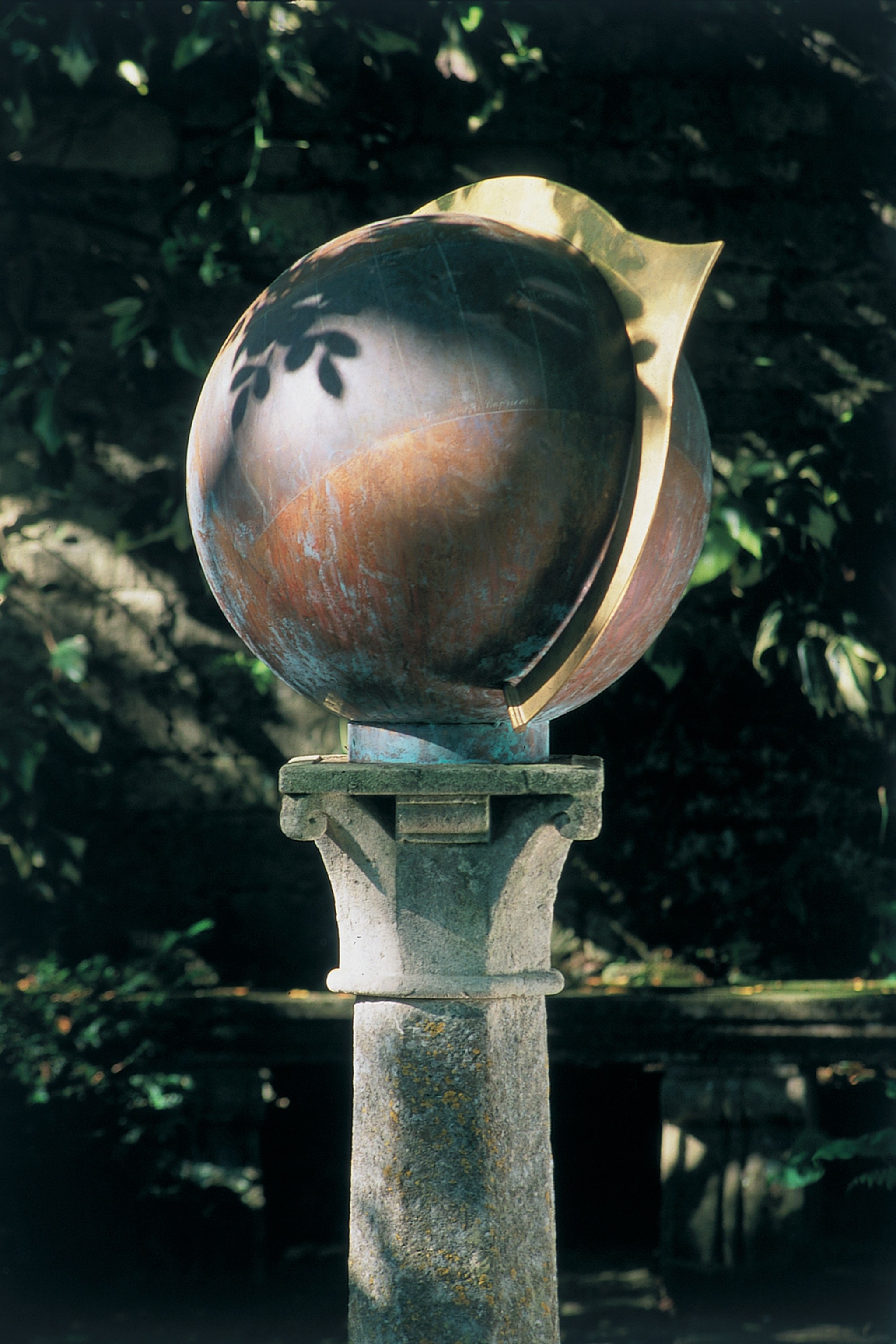 A globe copper sundial with a gold band sits on a stone plinth in a shaded garden.