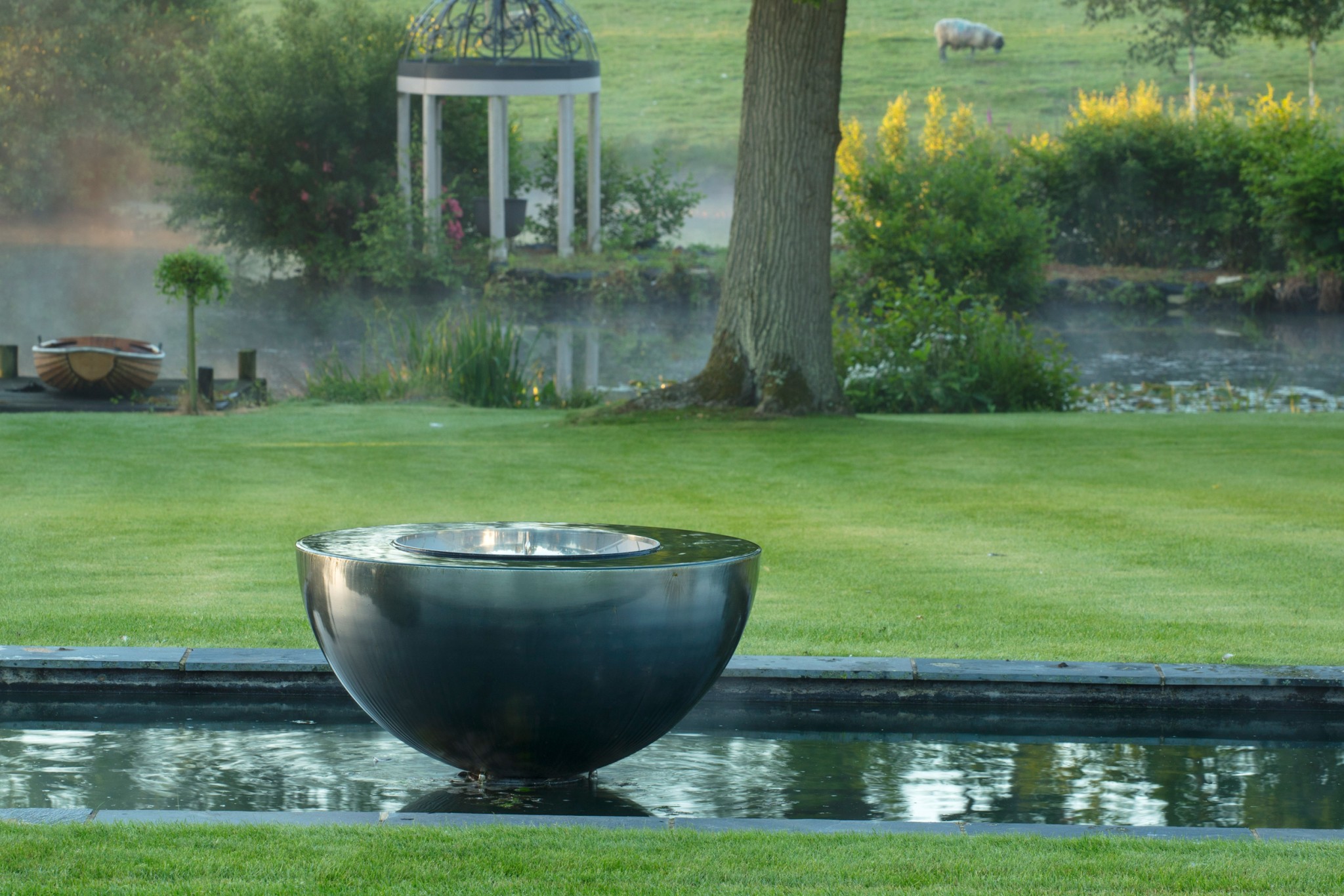 A stainless steel water chalice fountain gleams in a grassy garden, with a pond and gazebo behind this contemporary sculpture.