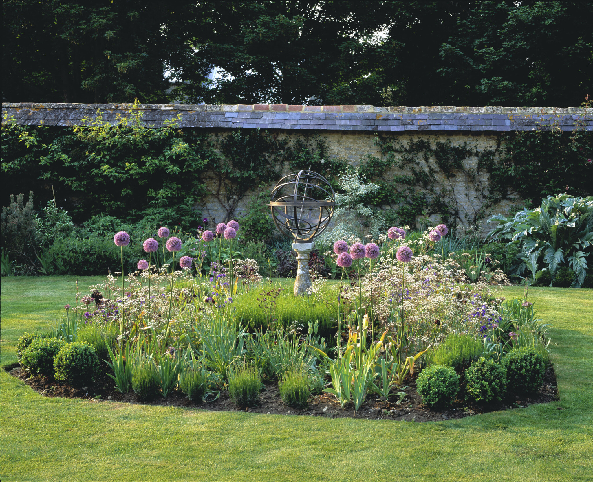 A garden with purple flowers and an armillary sphere sundial sculpture in front of a stone wall.