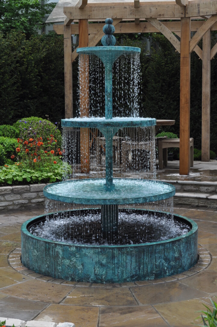 Three-tiered Crucello water fountain with cascading water, surrounded by stone paving and garden greenery.