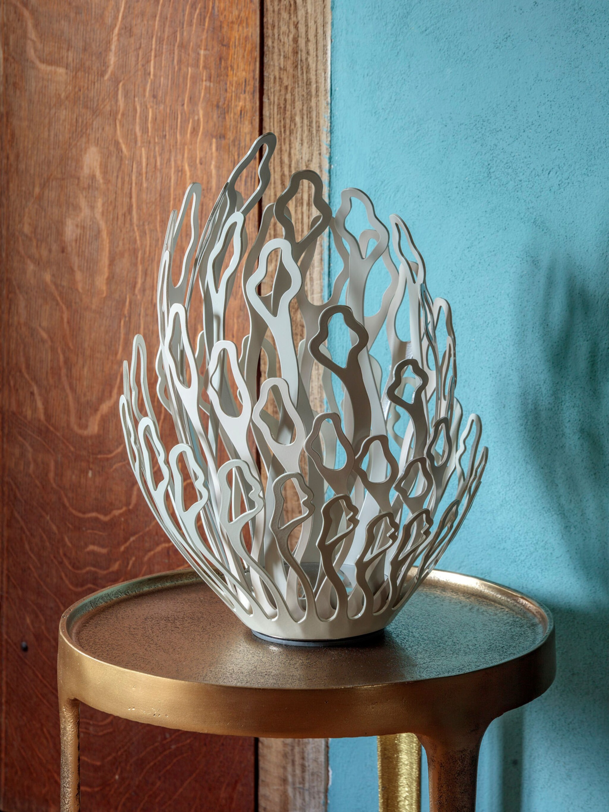 A white, abstract, metal sculpture resembling coral sits on a round, gold side table. The background features a turquoise textured wall and a wooden door. Soft natural light highlights the sculpture's intricate cut-out design.