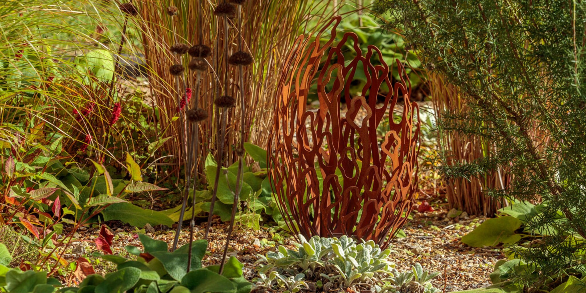 A rust-coloured, abstract metal sculpture shaped like an elongated egg stands among green and brown plants, grasses, and succulents on a bed of small pebbles in a sunlit garden.