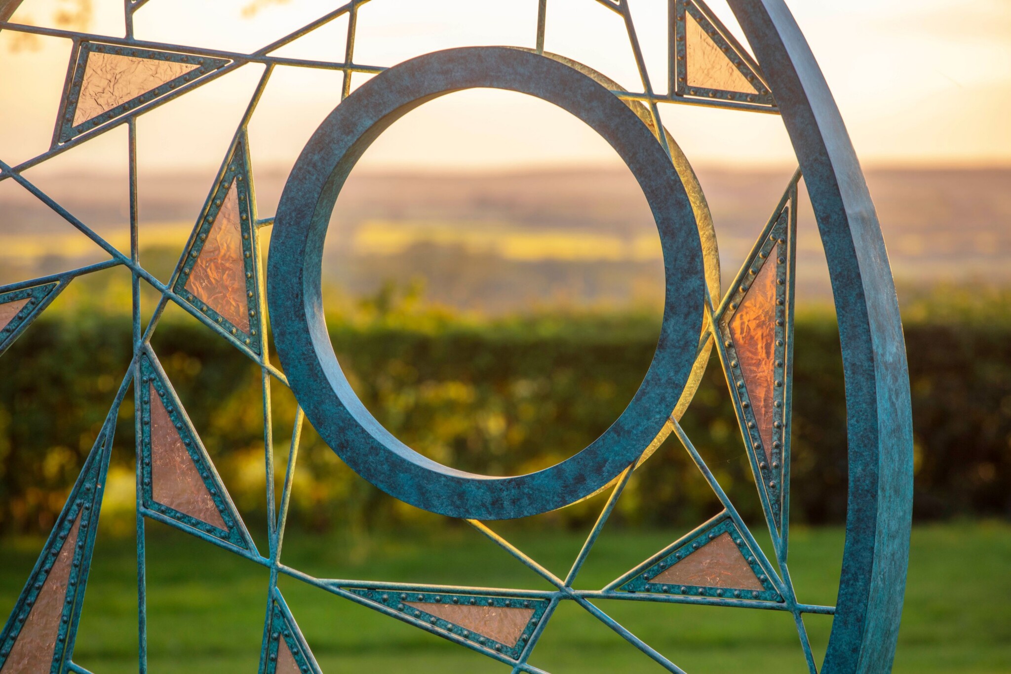 At sunset, the circular metal void of a bronze metal outdoor sculpture reveals a snapshot of the countryside in the distance. The triangles of copper mesh held between bronze rods glow in the fading light.
