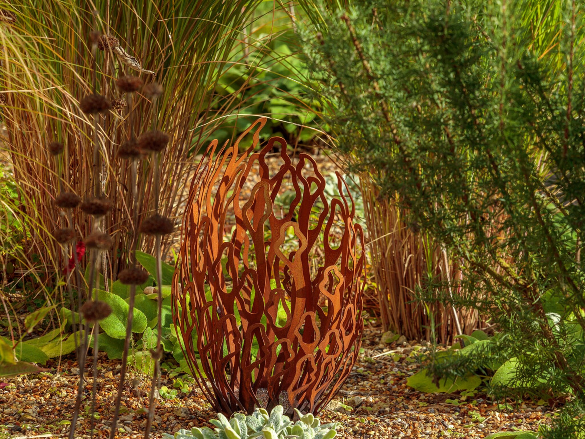 A rust-coloured metal garden sculpture with an abstract flame or leaf pattern stands among green and brown grasses, succulents, and textured plants in a landscaped garden with gravel ground cover.
