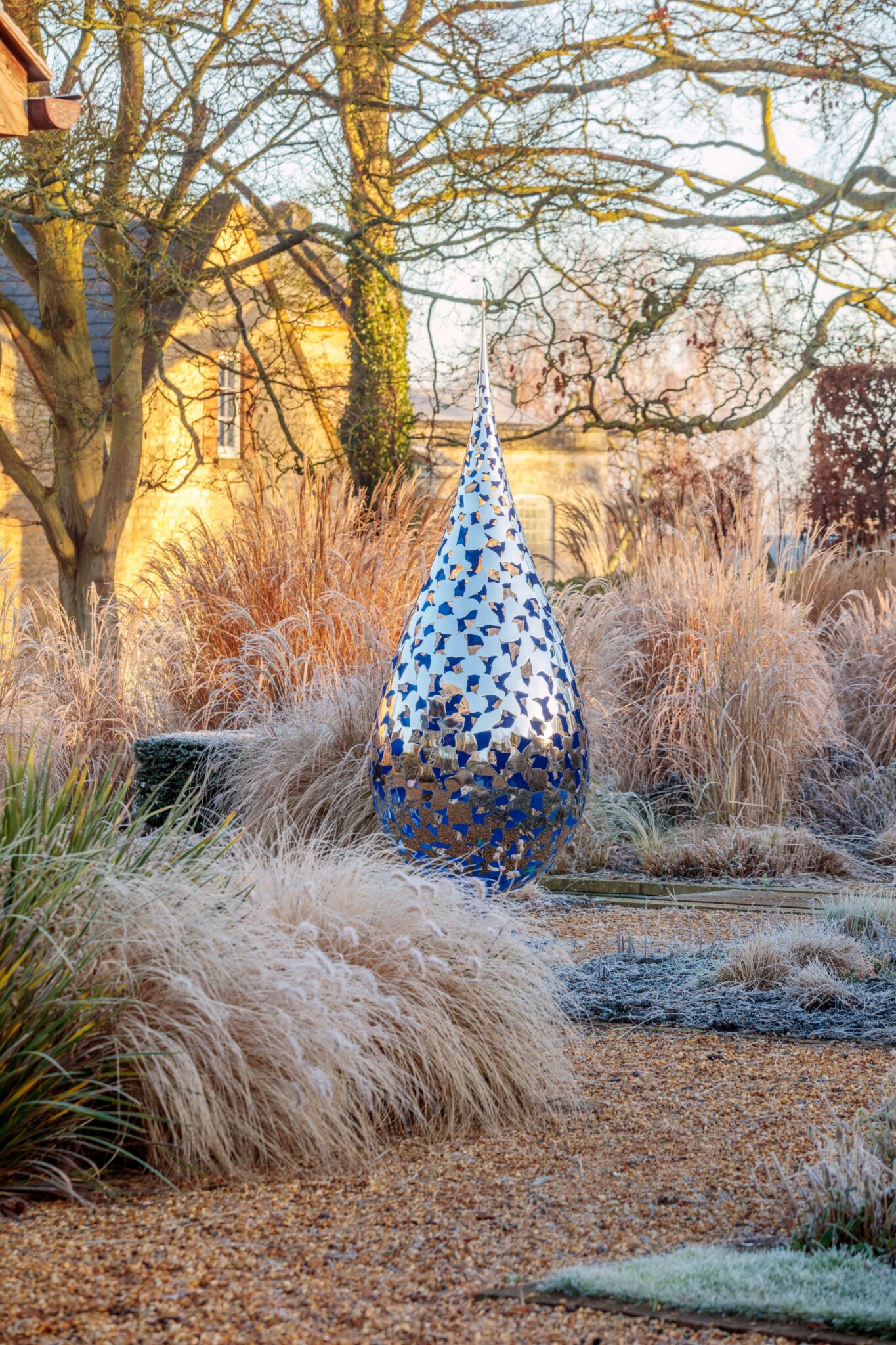 Shiny raindrop-shaped sculpture with delicate cut-outs in a frosty garden with tall grasses and trees.