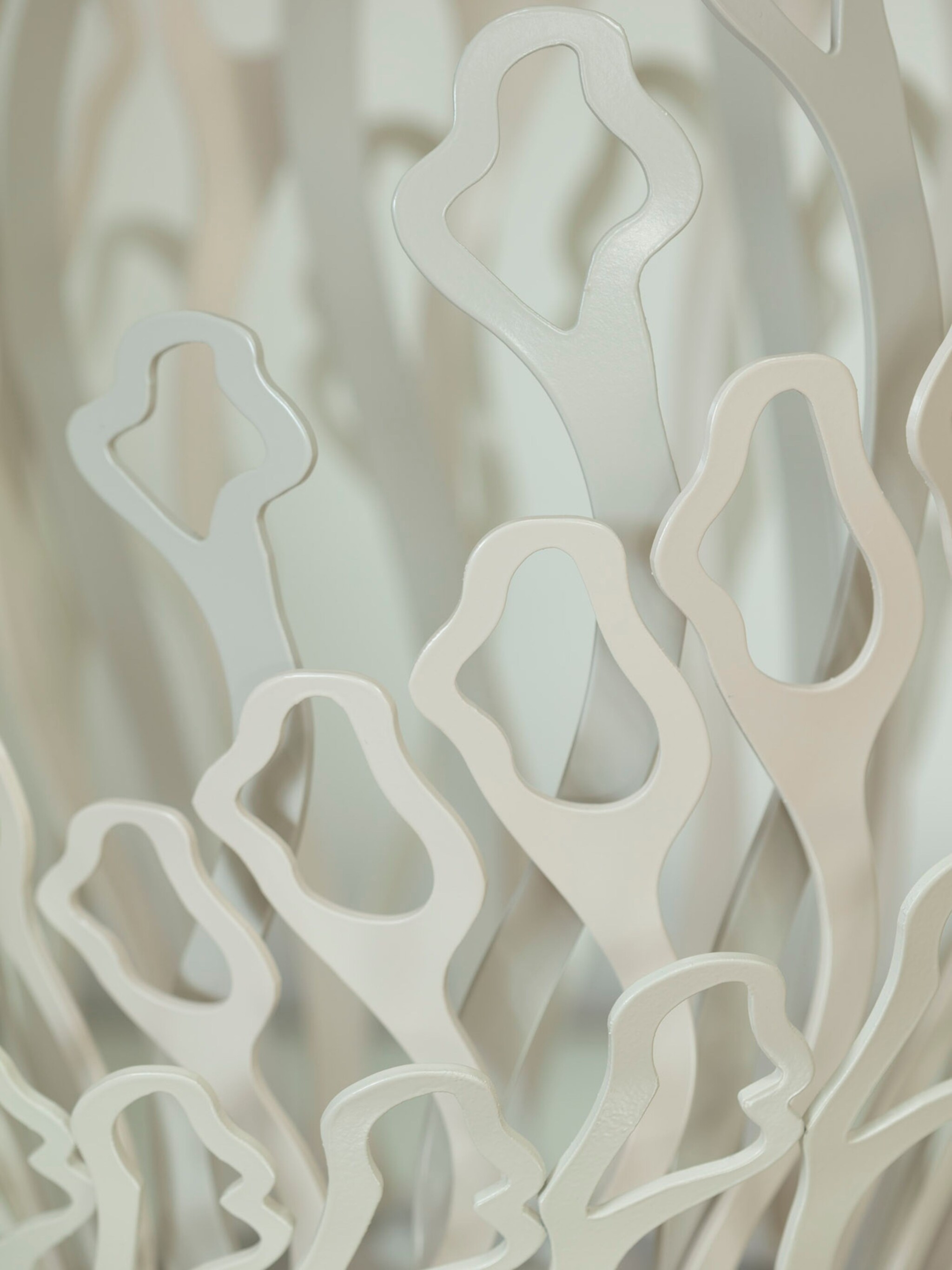 Close-up of overlapping, abstract metallic shapes with soft curves and cut-out centres. The pieces are a light, silvery colour and appear layered, creating a sense of depth and organic movement against a softly blurred background.
