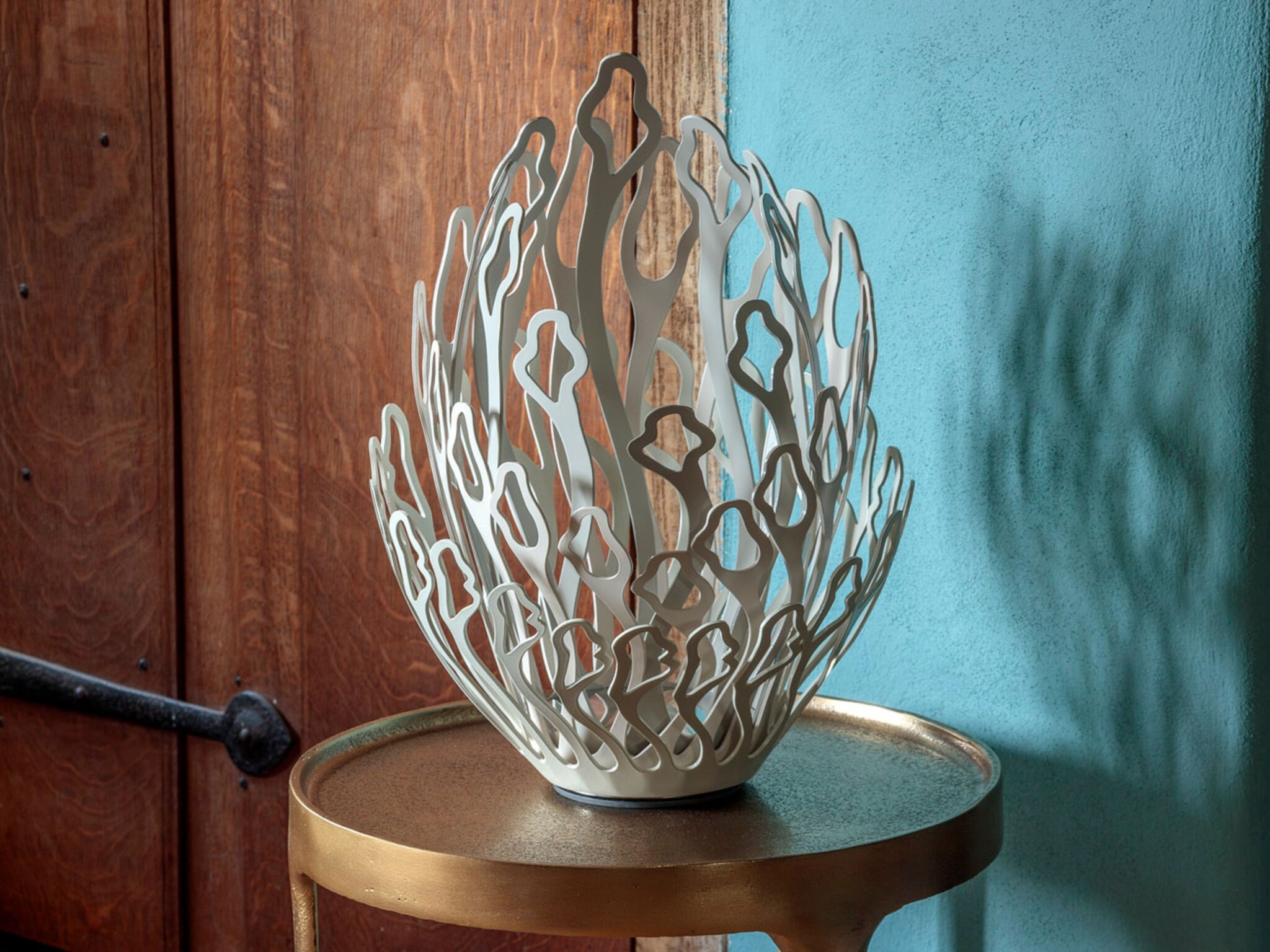 A sculptural white vase with an open, intricate, geometric cut-out design sits on a round, gold-coloured side table. The background features a wooden door and a blue textured wall, with the vase’s shadow cast onto the wall.