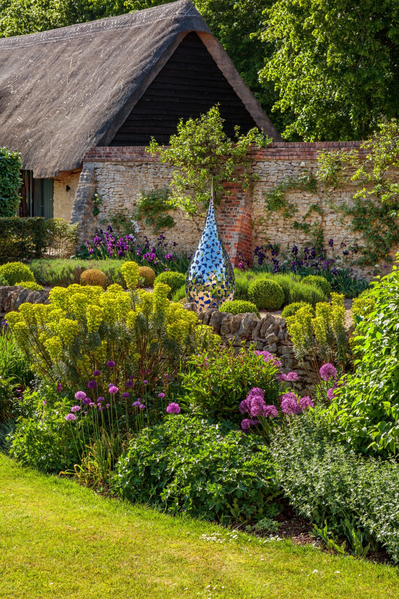 Colourful garden with flowers, a stone wall, a mosaic sculpture, and a thatched-roof building in the background.