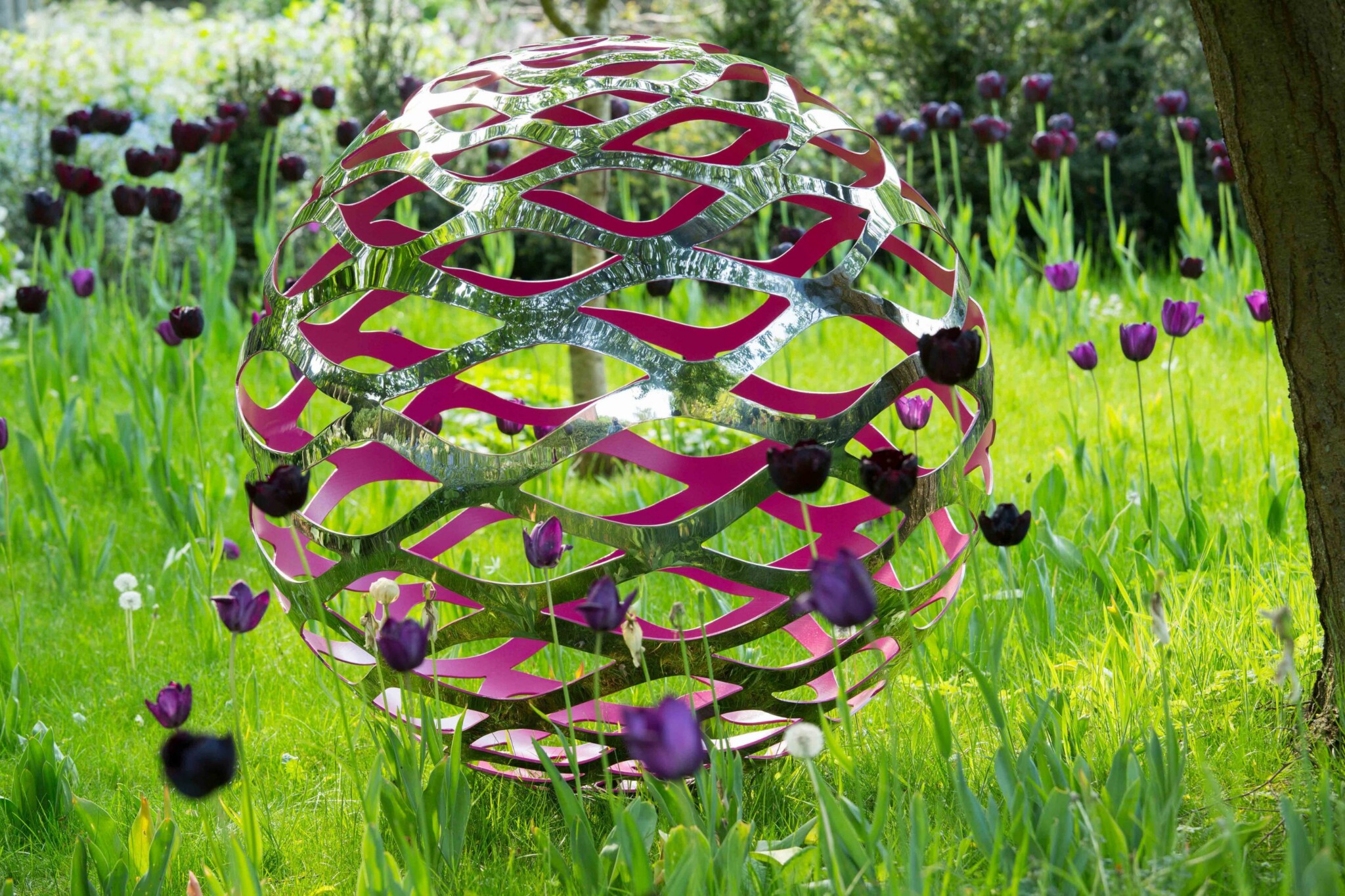 A shiny, spherical metal sculpture with pink accents sits among purple tulips in a grassy garden.