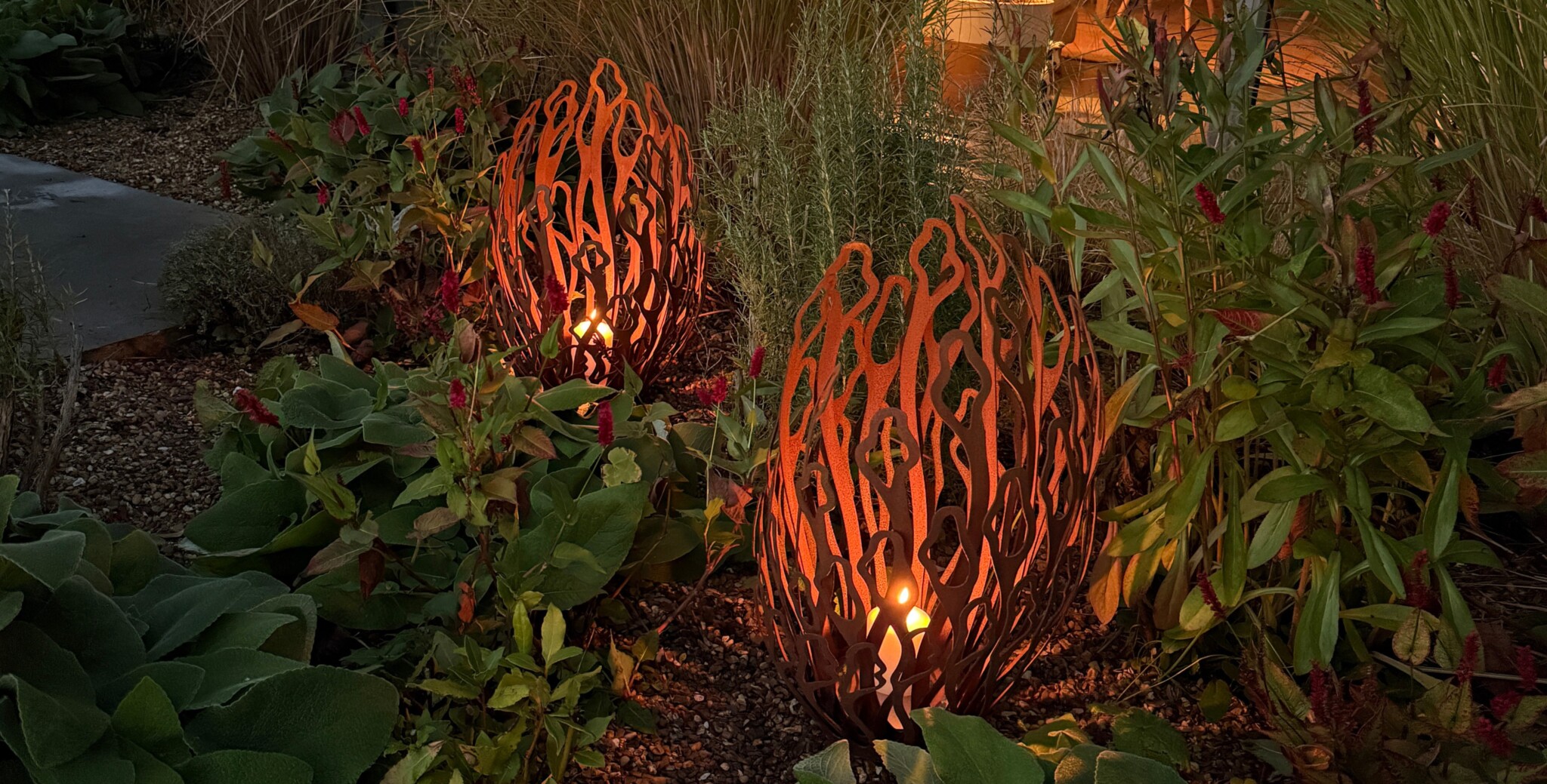 Two decorative metal lanterns cast a glow near an alba oxidised steel abstract sculpture in a leafy garden at dusk.