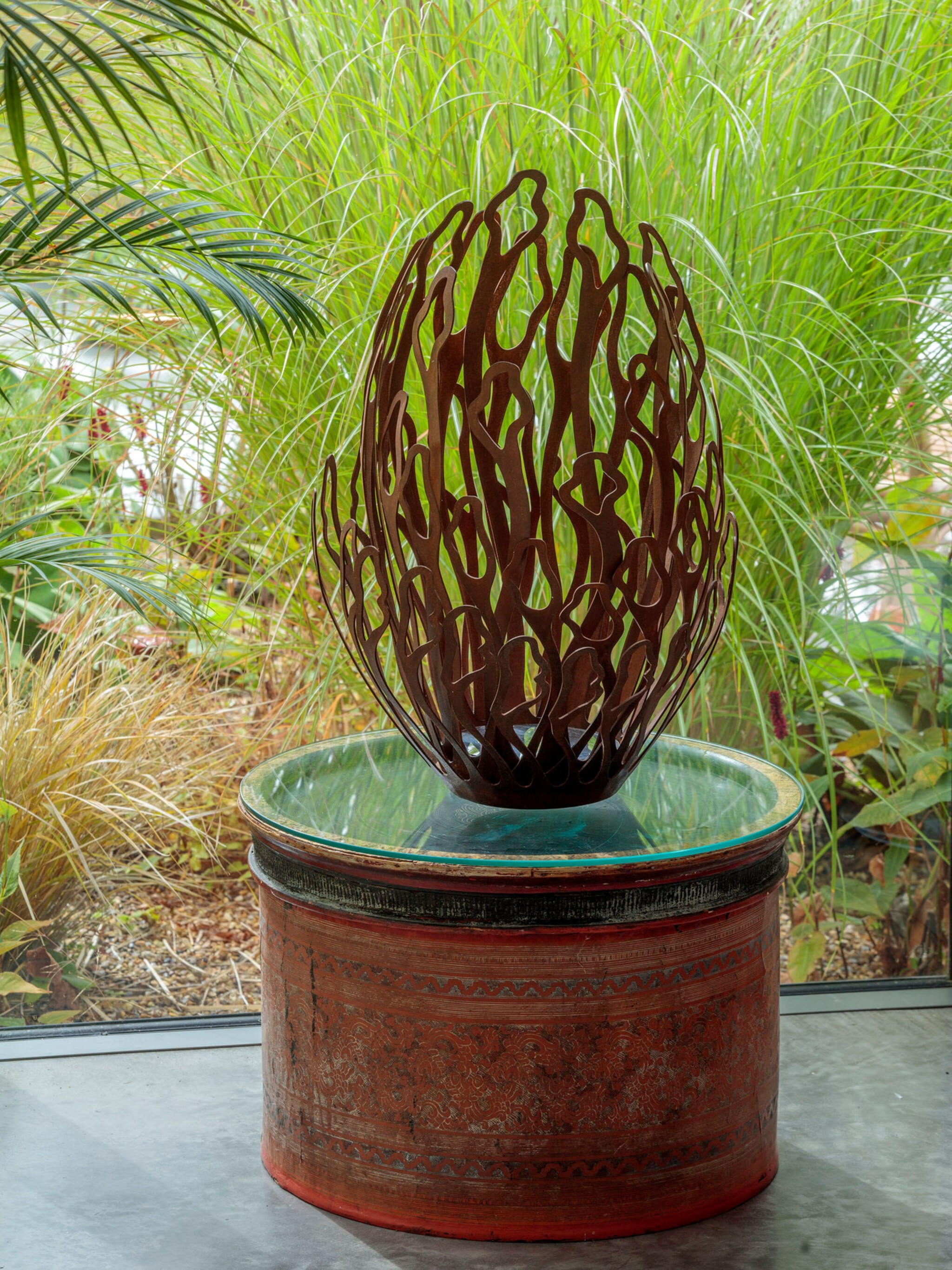 A decorative, abstract metal sculpture resembling stylised flames or leaves sits on a round glass-topped table with a patterned red base. Lush green grass and plants fill the background, suggesting an indoor garden or conservatory setting.