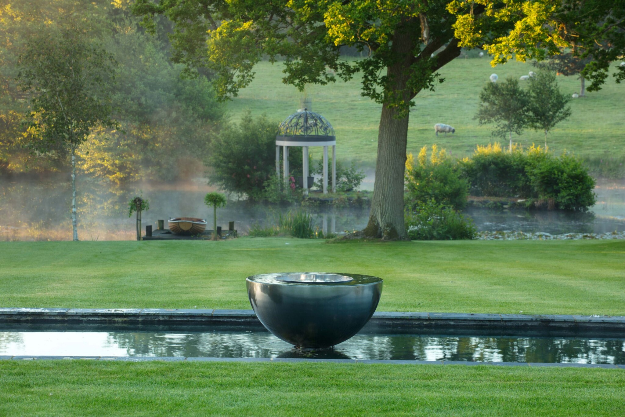 Stainless steel bowl water feature installed above formal pool in an English landscape garden.