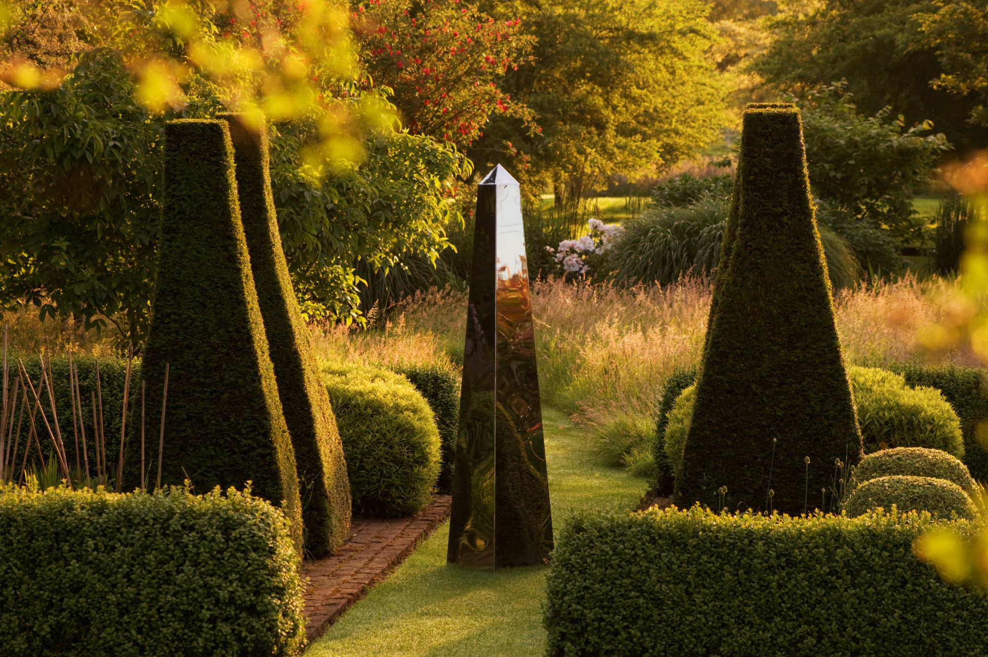 A reflective obelisk stainless steel sundial stands between two tall hedges in a lush, sunlit garden.