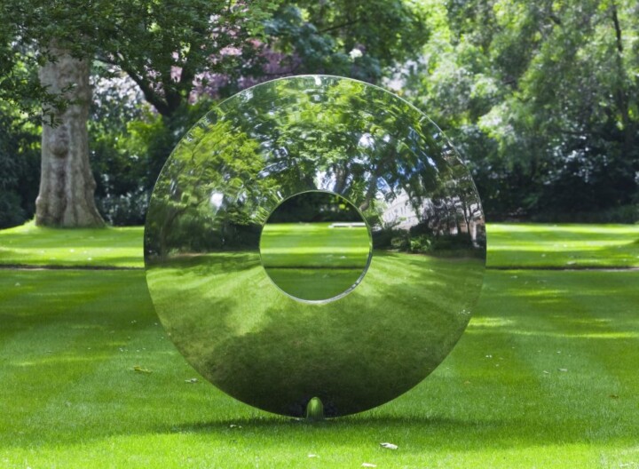 A large, shiny torus stainless steel contemporary abstract sculpture with a central hole stands on a manicured green lawn. Its polished surface mirrors the surrounding trees and sunlight, blending seamlessly into its lush park setting.