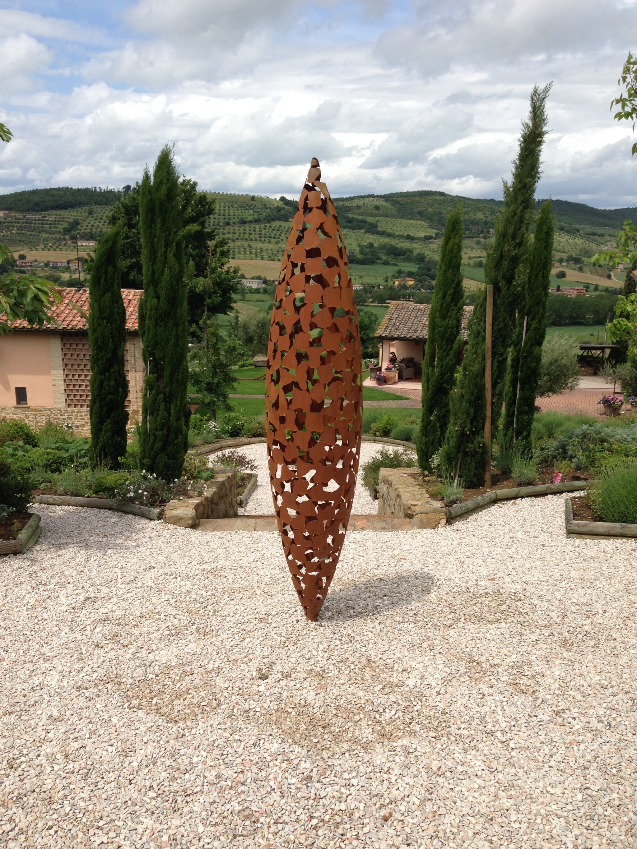 A column-like sculpture in chestnut-hued oxidised steel stands sentry on a gravel courtyard with pencil pines in the background.