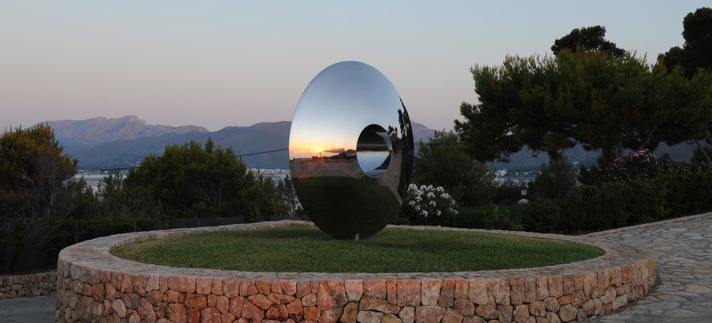 A contemporary abstract torus sculpture of shiny stainless steel with a central hole stands on a grass patch bordered by stone. The sculpture reflects the sunset, trees, and mountains as a stone path curves beside it, surrounded by shrubs and trees.