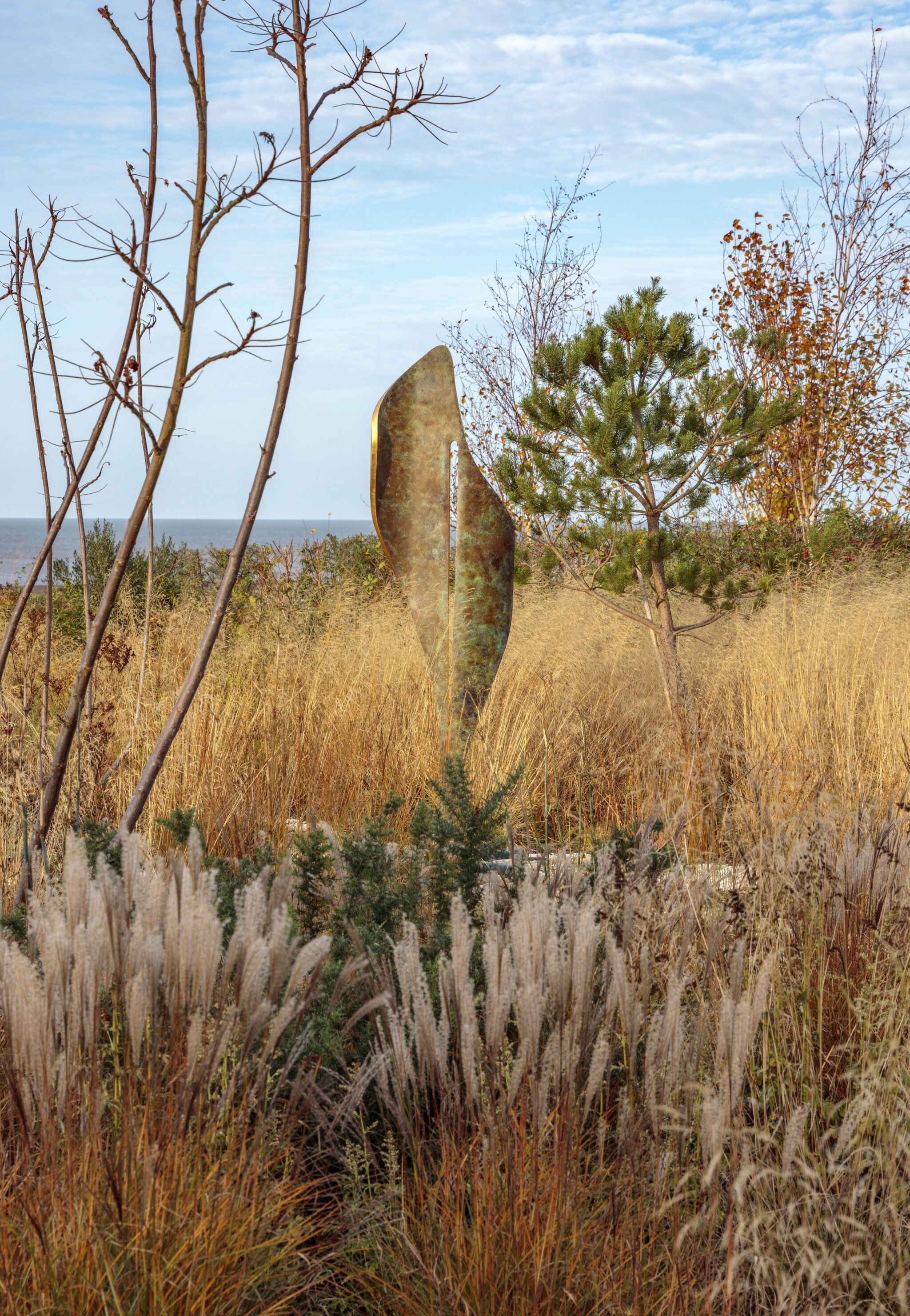 Abstract Florio sculpture made from verdigris copper patina and 24-carat gold leaf stands among tall grasses near the water.