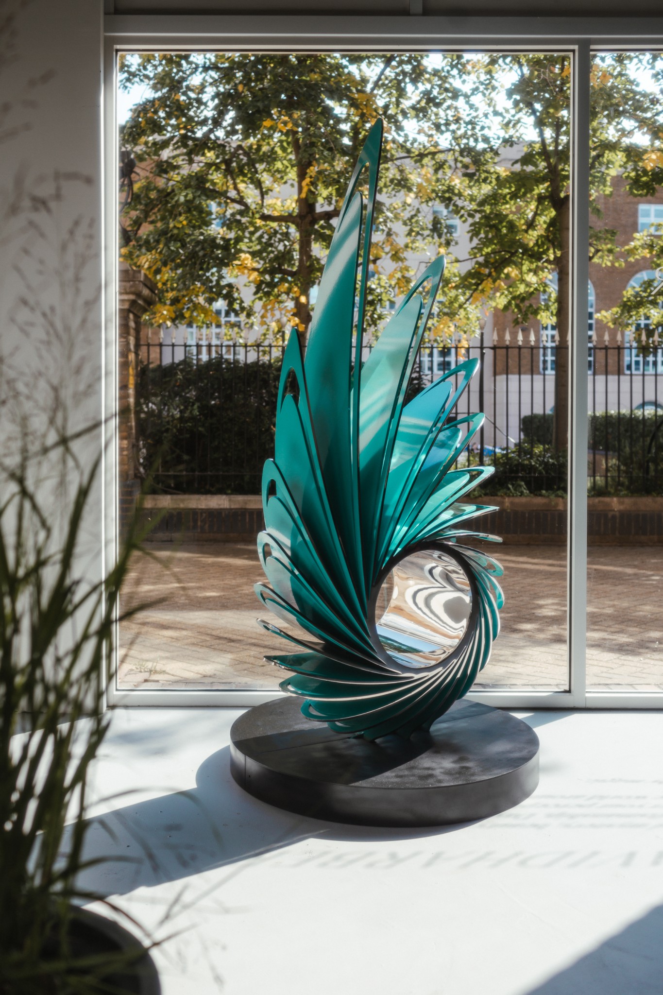 A modern, teal glass and Orbis modern metal sculpture with wing-like curves stands by a sunlit window.