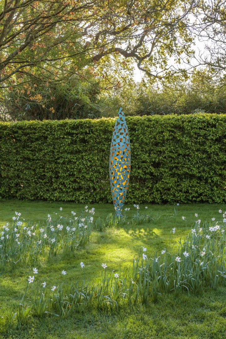 Colorful quill bronze and gold leaf contemporary abstract sculpture stands on green lawn with white flowers and trees in another angle
