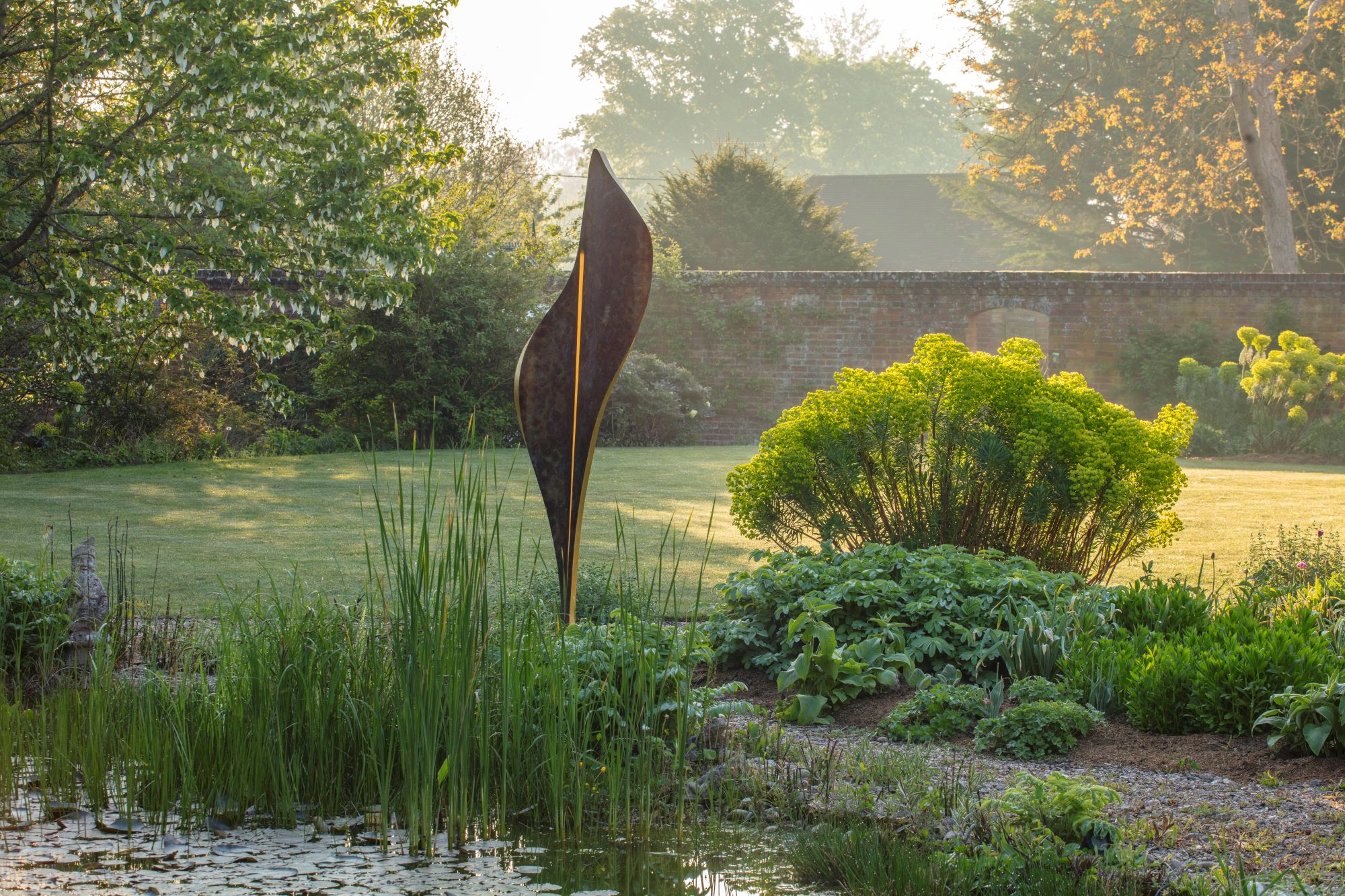 A tranquil garden scene at sunrise features a tall Flo Contemporary Abstracts sculpture made from bronze finish and gold leaf near lush green plants by a pond. Sunlight gently illuminates the landscape, creating a peaceful atmosphere.