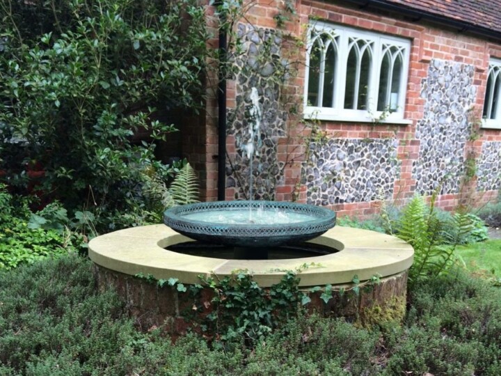 A round bird bath sits on a stone pedestal near a Mimeo bronze abstract sculpture, with a brick house in the background.