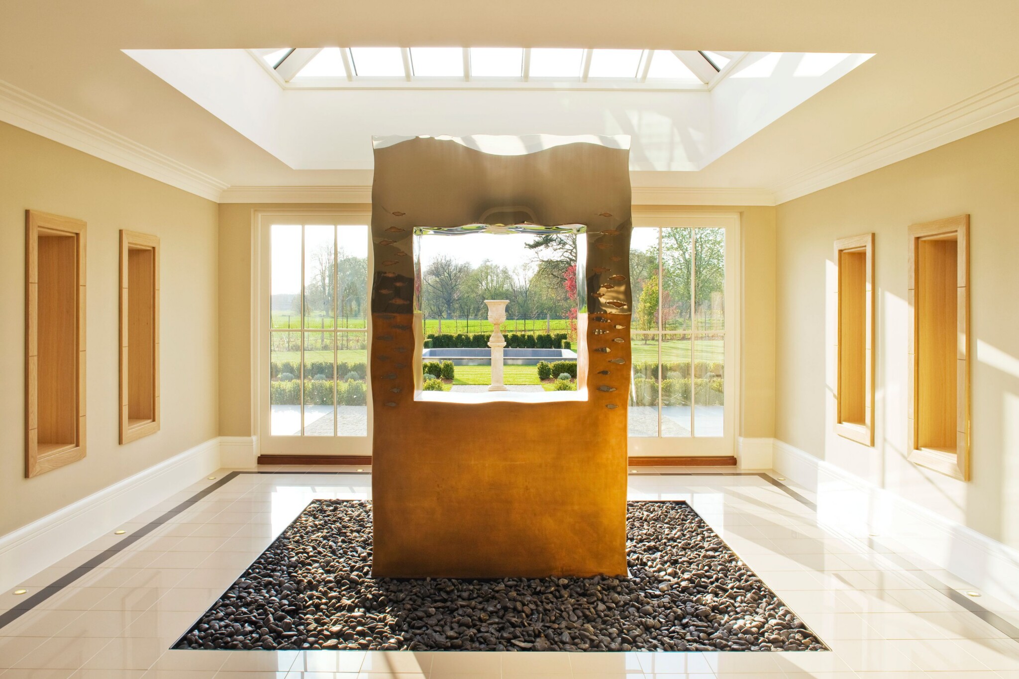Corten Steel metallic frame, perfect for hallway sculpture ideas, stands on stones; urn and green trees seen through windows behind.