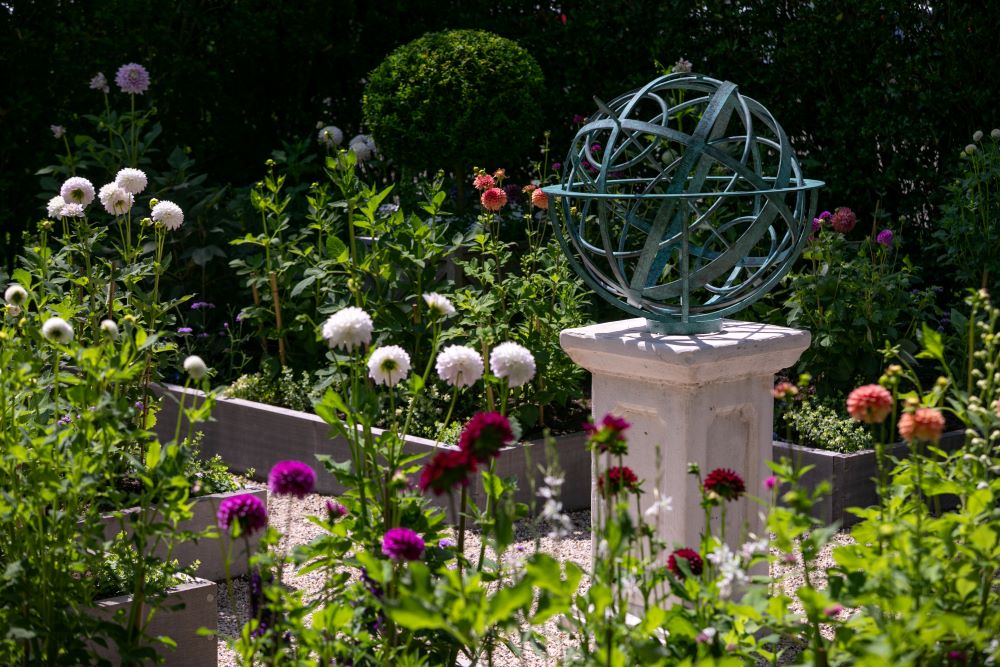 A bronze verdigris armillary sphere sculpture sits atop a white pedestal in a lush garden at East Hampton, New York, surrounded by blooming flowers and raised garden beds for a touch of contemporary elegance.